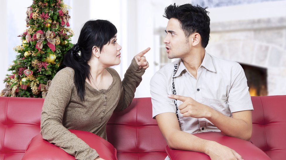 A seated couple argue in front of a Christmas tree